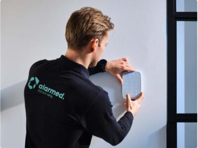 Persoon in zwart Alarmed-shirt bevestigt een slim alarmsysteem aan een witte muur naast een raam.